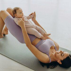 Une maman allongée sur un tapis de yoga pratique une posture avec son bébé allongé sur son ventre, dans un moment de partage et de mouvement doux.