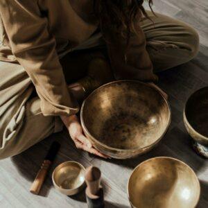 Personne assise au sol tenant un grand bol tibétain, entourée d'autres instruments sonores utilisés pour un bain sonore.