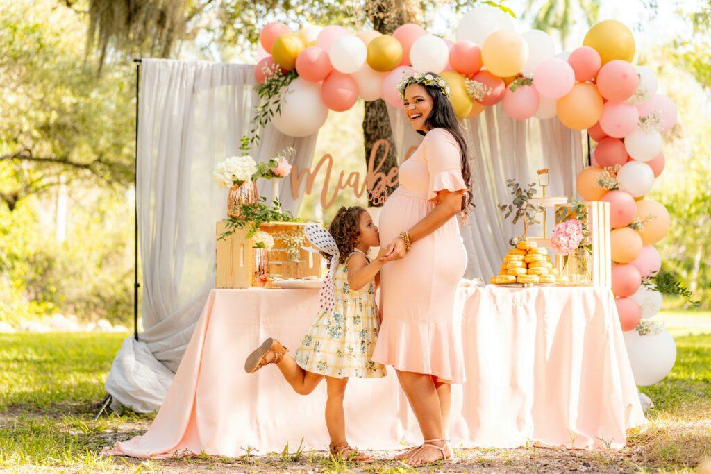Une femme enceinte souriante, vêtue d’une robe rose pâle et portant une couronne de fleurs, se tient devant une table décorée pour une fête en plein air. Une petite fille l'embrasse tendrement sur le ventre. La scène est ornée de ballons roses, blancs, dorés, et d’une décoration florale.