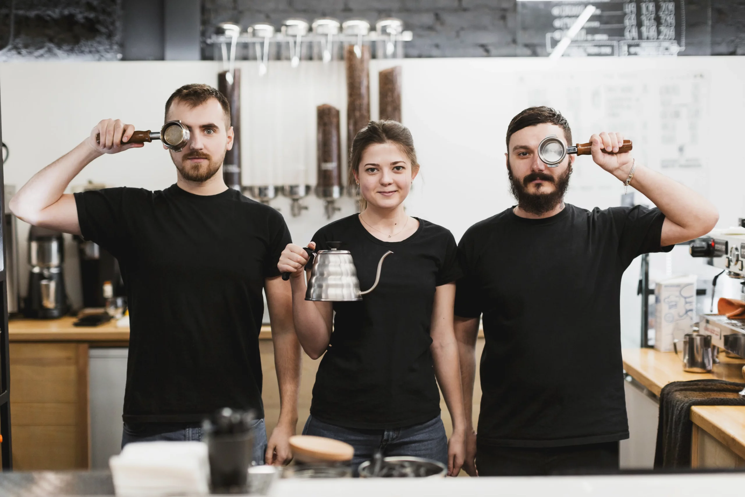 Trois baristas de Coffeart posent derrière le comptoir, tenant un porte-filtre et une bouilloire à col de cygne, symboles de leur passion pour le café et la création.