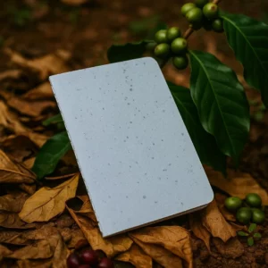 Carnet artisanal gris posé sur un lit de feuilles et grains de café verts, illustrant le papier recyclé enrichi au marc de café.