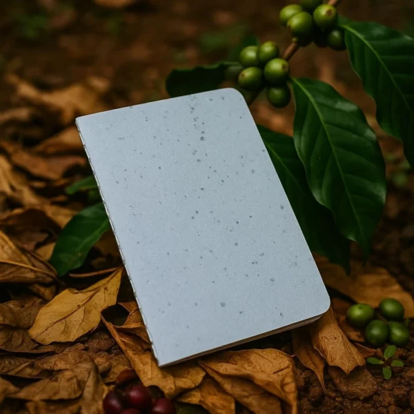 Carnet artisanal gris posé sur un lit de feuilles et grains de café verts, illustrant le papier recyclé enrichi au marc de café.