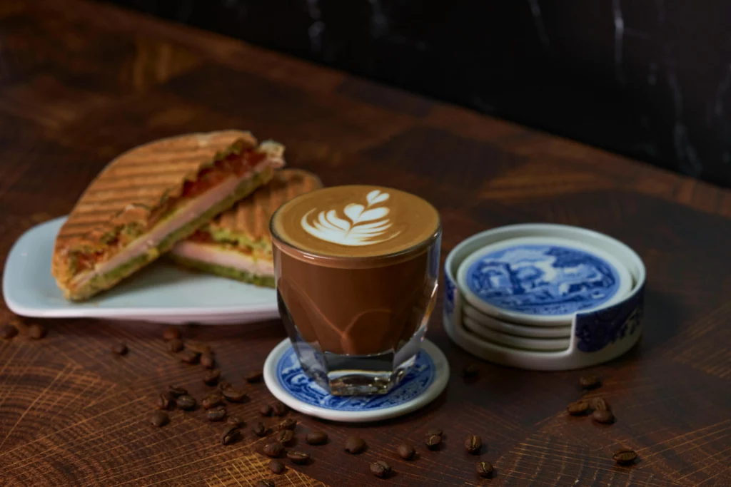 Gros plan d’un latte art dans un verre posé sur une soucoupe bleue, accompagné d’un panini grillé garni de fromage et légumes, sur une table en bois.
