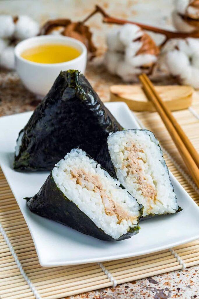 Deux onigiri, un entier et un autre couper en deux où on voit la garniture à l'intérieur. Ils sont dans une petite assiette rectangulaire sur une nappe en bambou avec des baguettes à coté