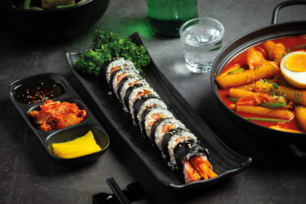 Kimbap présenter dans une petite assiette rectangulaire et coupé en plusieurs bout, il y a des petits accompagnements à coté avec du kimchi et du radis jaune. Il y a aussi un bout de plat de tteokbokki et un petit verre