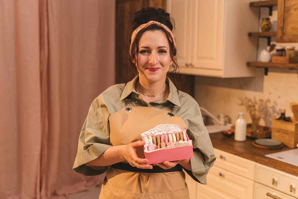Cheerful woman in apron presenting a box of colorful macarons in a cozy kitchen.
