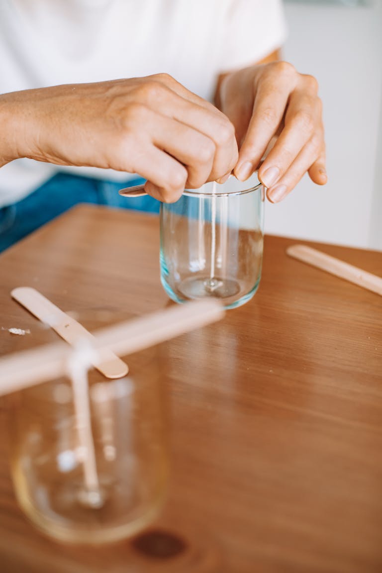 Hands working on a glass candle crafting project with wooden sticks.