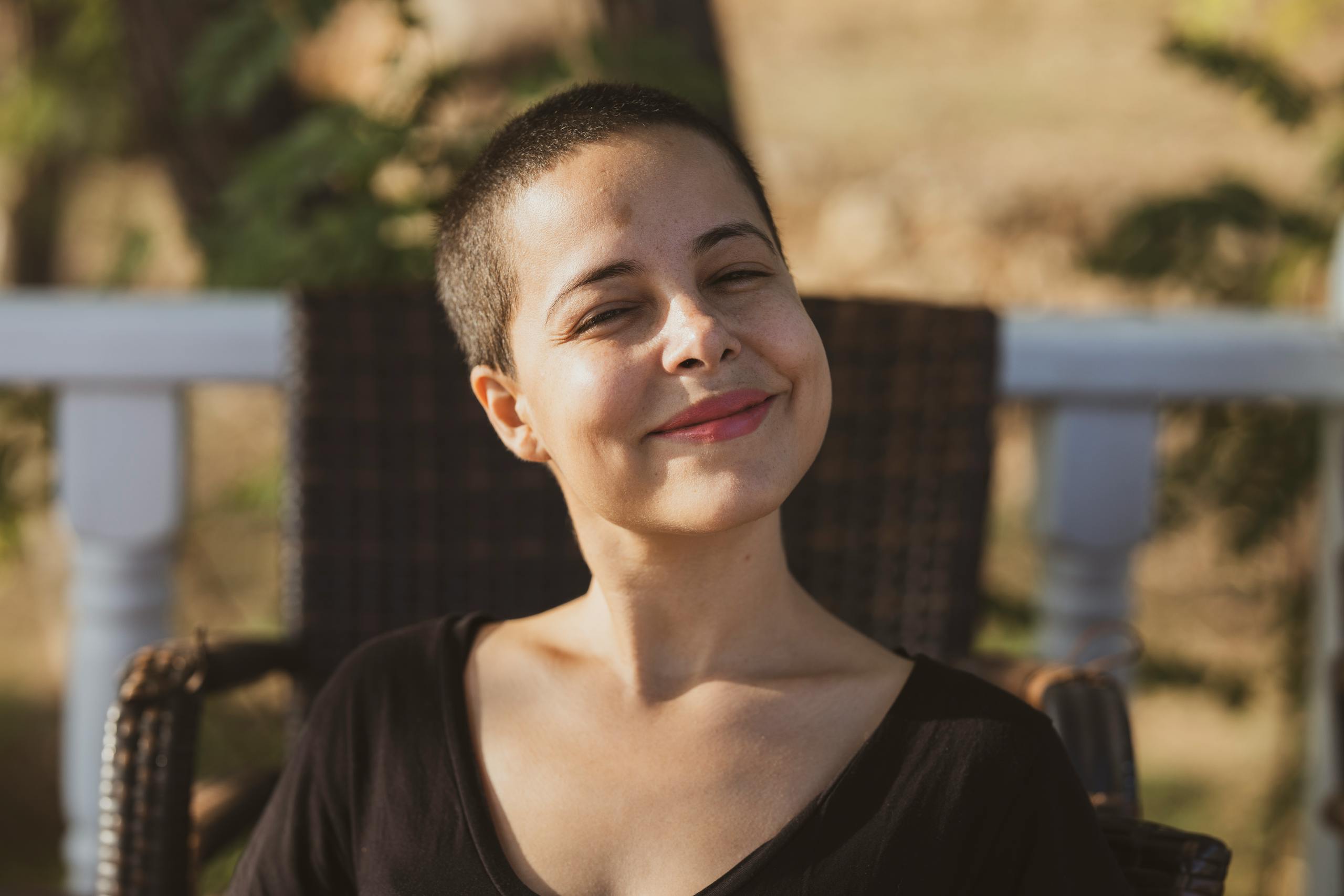Femme aux cheveux rasés assise au soleil, sur un siège, souriante.