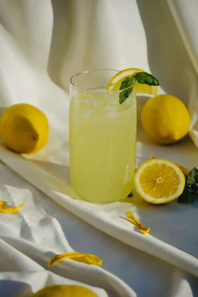 Refreshing lemonade with mint and ice surrounded by whole lemons on white draped fabric.