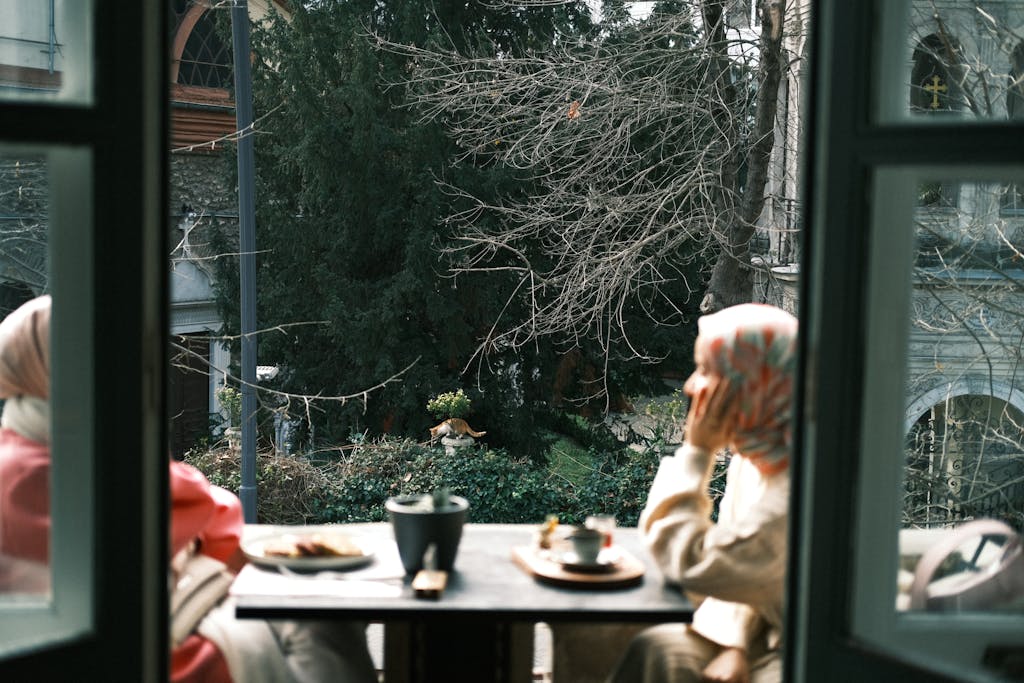 Two women enjoy coffee on a cozy balcony, surrounded by urban greenery.