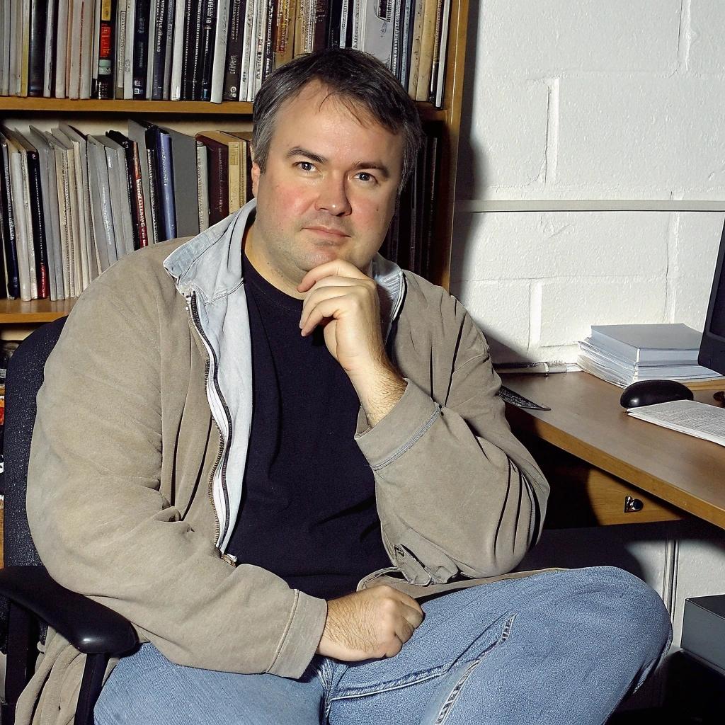 homme assis sur une chaise de bureau avec gilet marron et t-shirt bleu foncé. Devant une bibliothèque.
